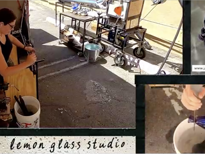Sample image of a twitch stream, there are two camera angles showing a woman making a hand blown cobalt blue glass. One camera focuses on her hands, while she uses the jacks to create a constriction where she will make a clean break to define the lip of the cup. Also in frame is a small purple glass octopus in a top hat, and Lemon Glass Studio Logo